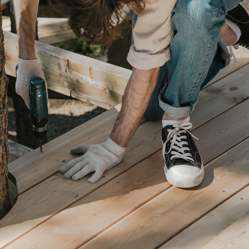 Worker in white gloves driving a screw into new wooden deck boards with a cordless drill