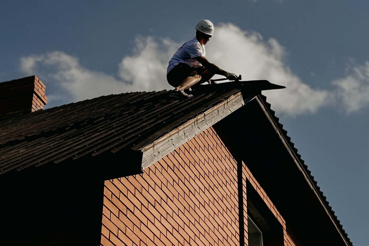 Roofer crouched at the ridge of a steep roof on a red brick house against a blue sky