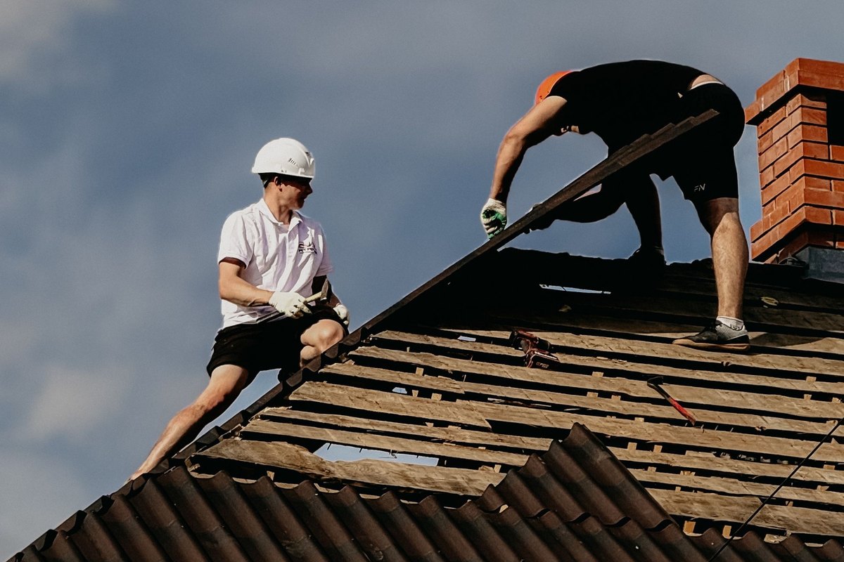 Two roofers working on a steep dark roof with exposed wooden battens near a brick chimney