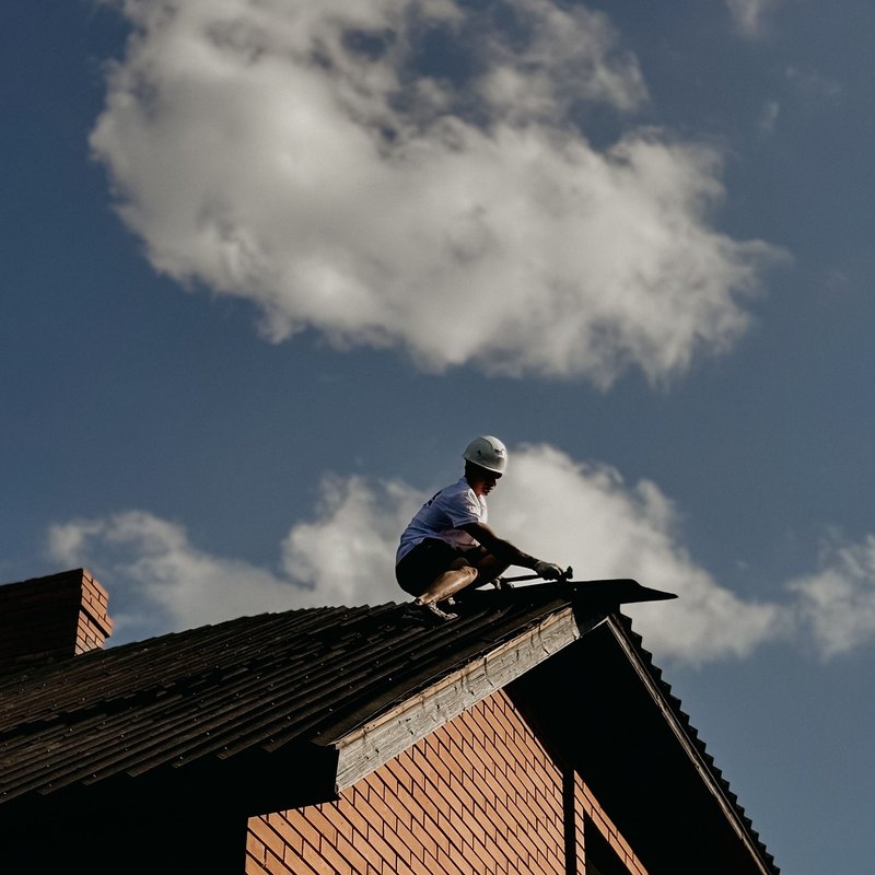 Roofer crouched at the ridge of a steep roof on a red brick house against a blue sky