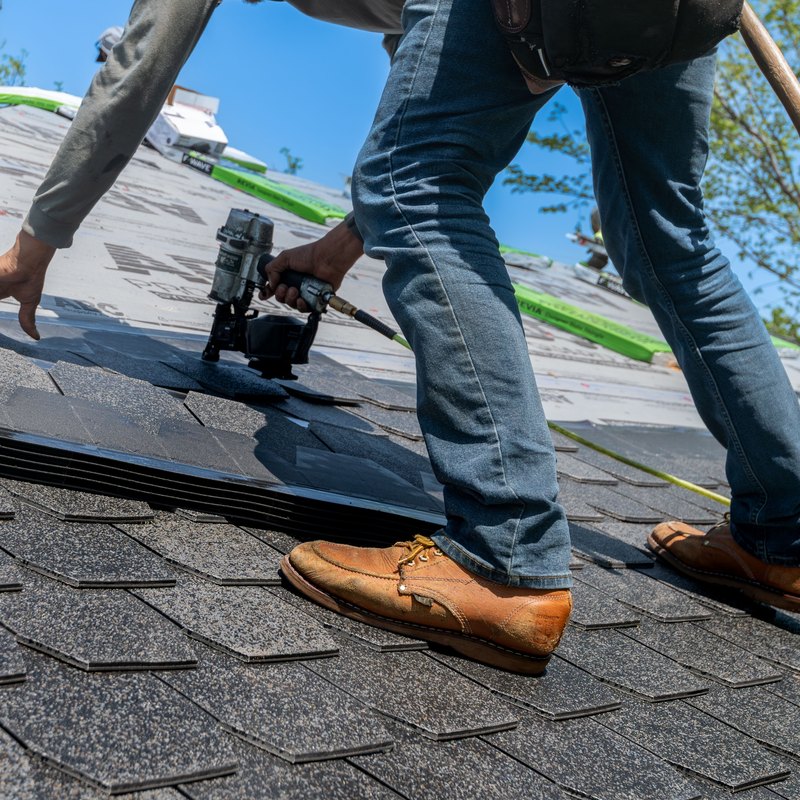 Roofer with a nail gun installing gray asphalt shingles on a sloped roof under a blue sky