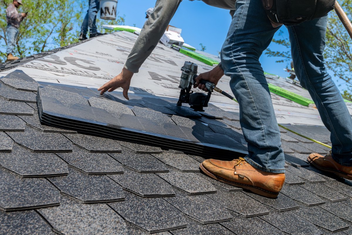 Roofer with a nail gun installing gray asphalt shingles on a sloped roof under a blue sky