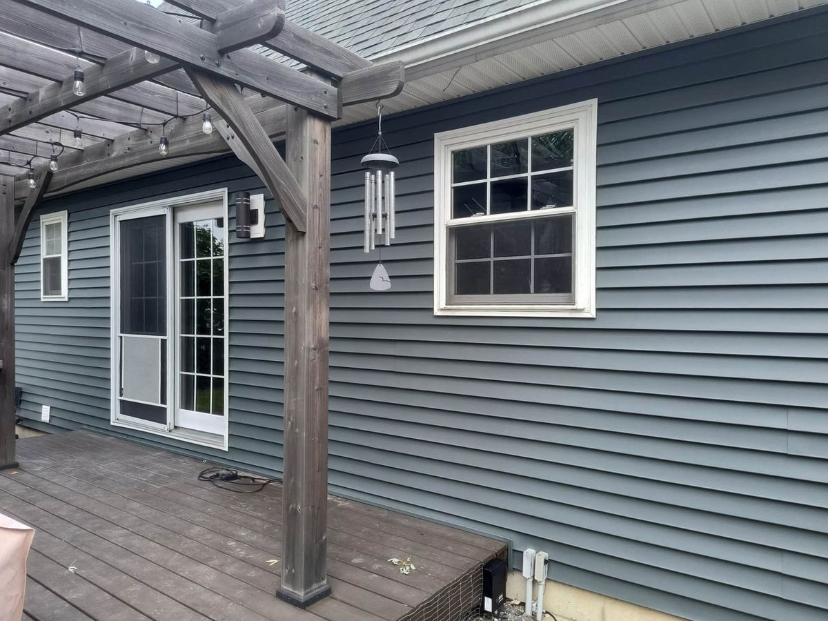 Rear wall of a house in slate blue-gray vinyl siding with a wood pergola over a brown deck and a double-hung window