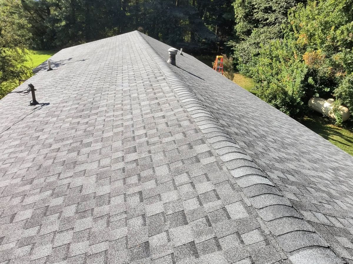 Finished gray asphalt shingle roof with a capped hip, plumbing vents and a stepladder on the lawn beside it