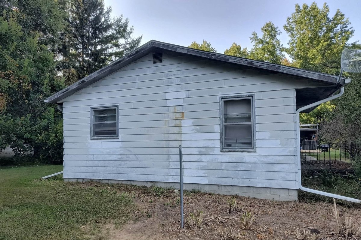 Weathered white lap siding patched with primer on an older one-story house with two old windows