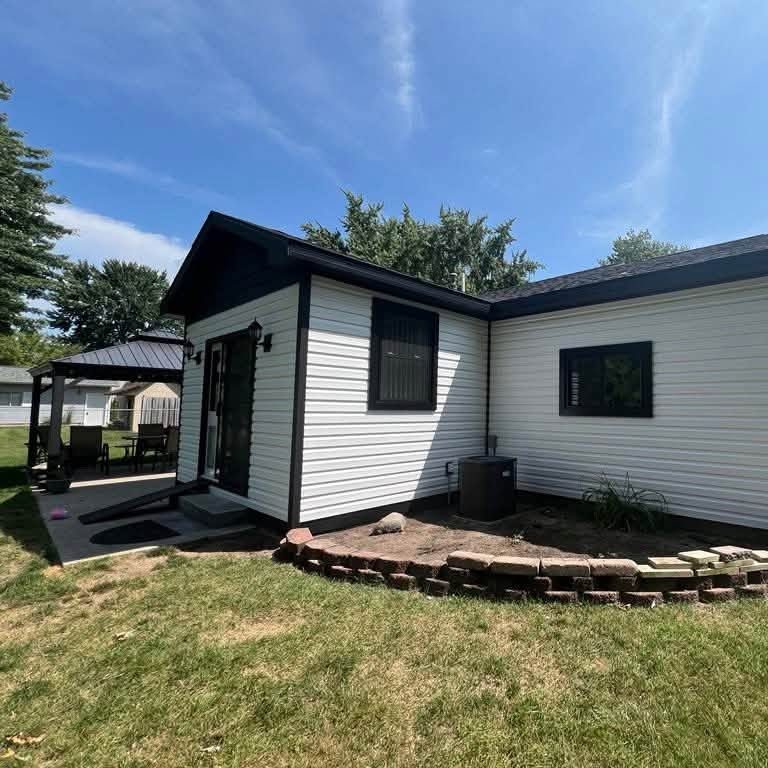 White horizontal vinyl siding on a small house with black trim, a glass door and a gazebo at the left