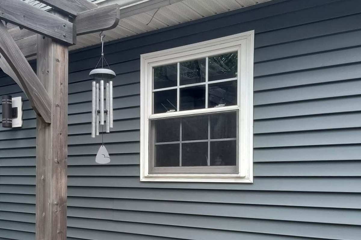 Rear wall of a house in slate blue-gray vinyl siding with a wood pergola over a brown deck and a double-hung window